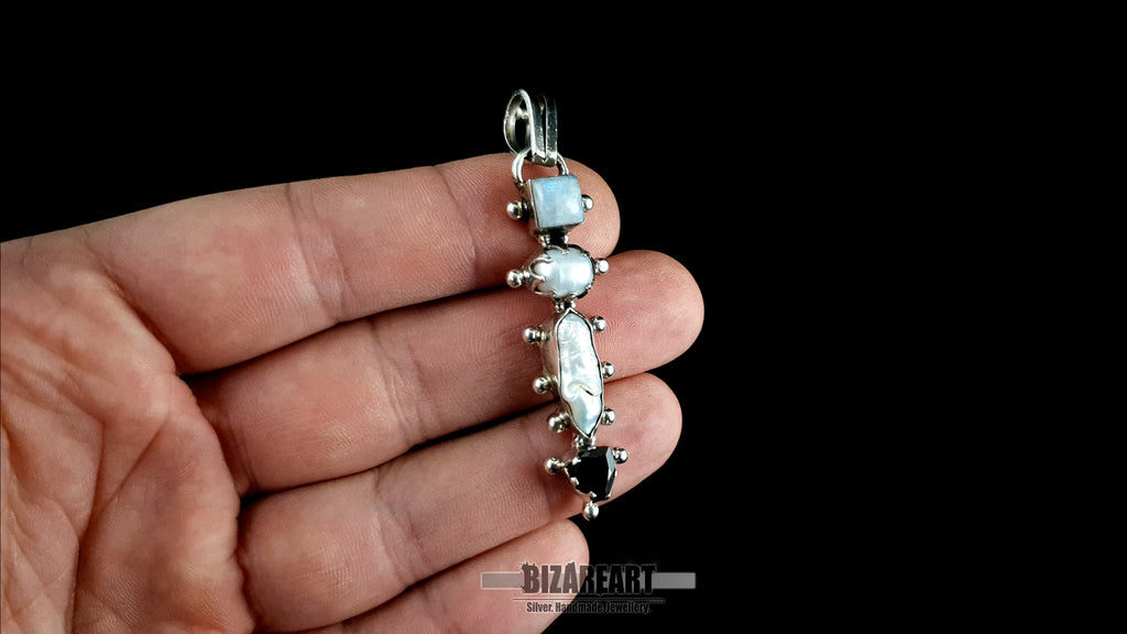 A close-up image showing the intricate craftsmanship of the sterling silver multi-gemstone pendant as it is carefully held and displayed in a person's hand, revealing the detailed settings and natural variations of the moonstone, pearl, and garnet.