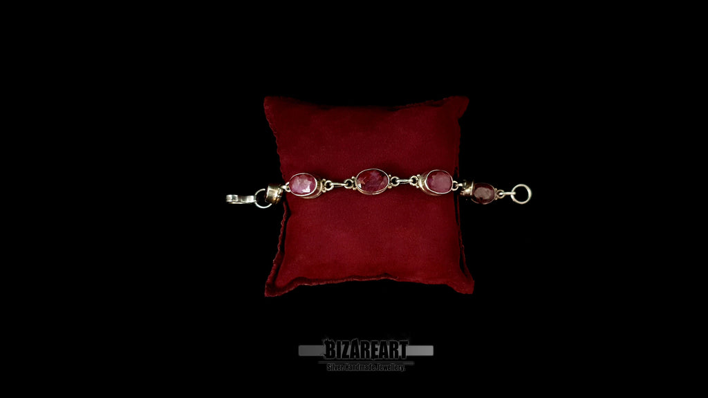 Studio flat lay of a .925 sterling silver artisan link bracelet with six red ruby stones on a red velvet display pillow.