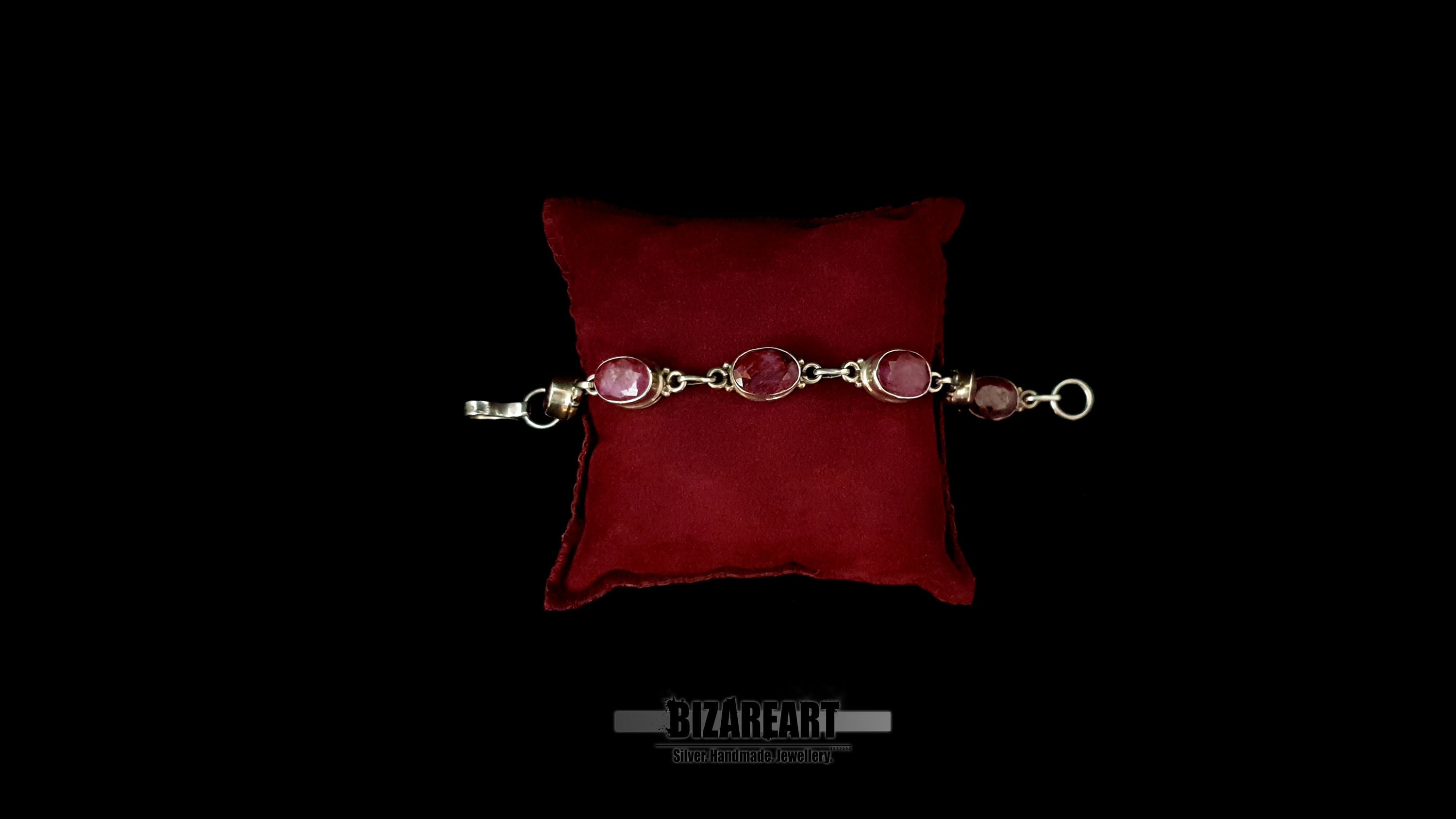 Studio flat lay of a .925 sterling silver artisan link bracelet with six red ruby stones on a red velvet display pillow.