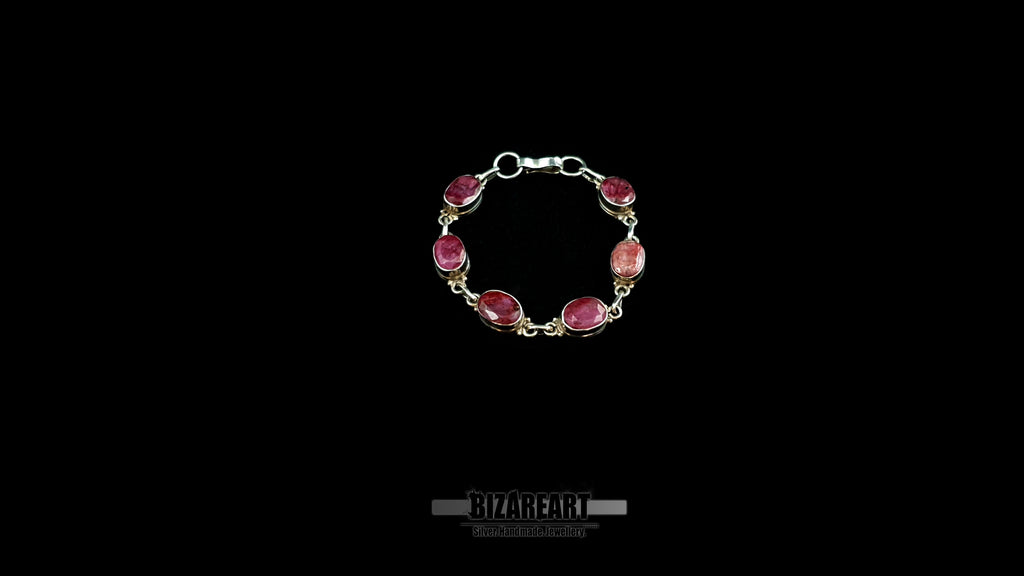 Centered product shot of an open-clasp sterling silver bracelet set with six faceted raw pinkish-red ruby gemstones.