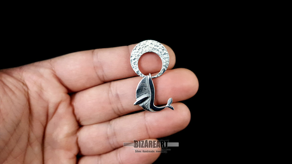 Close-up of a hand holding the kinetic sterling silver whale and moon pendant to show scale. The image highlights the intricate hand-engraved details on the whale's body and the unique textured surface of the hammered silver moon.