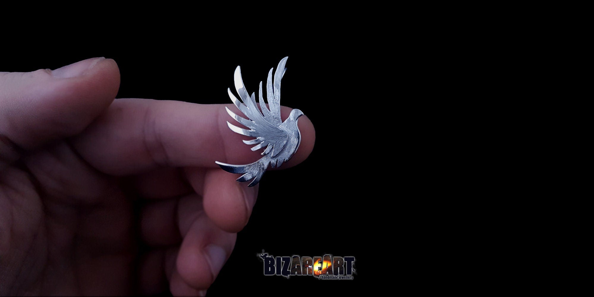 A close-up photo of the Bird pendant held between two fingers to demonstrate its physical size and the fine details of the silver craftsmanship.