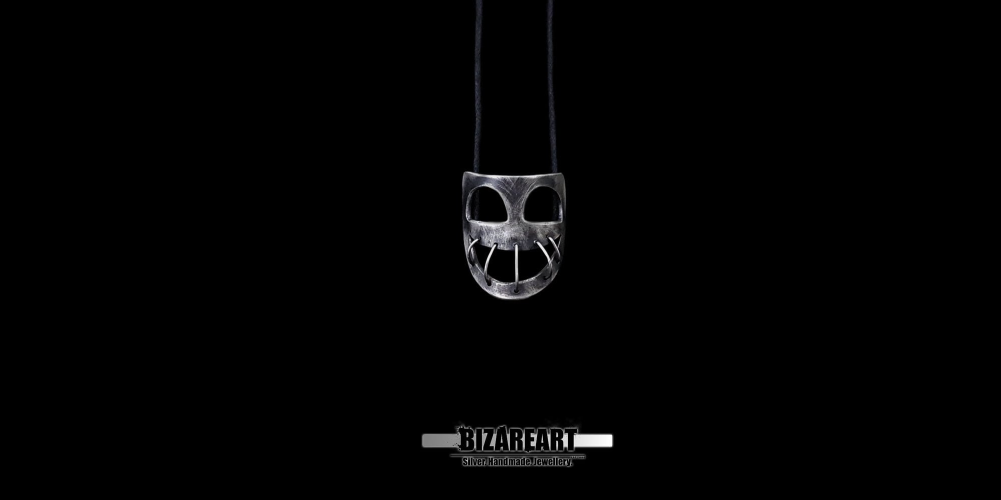 Distant, minimal shot highlighting the eerie, dark elegance of the small silver mask necklace against a black backdrop.