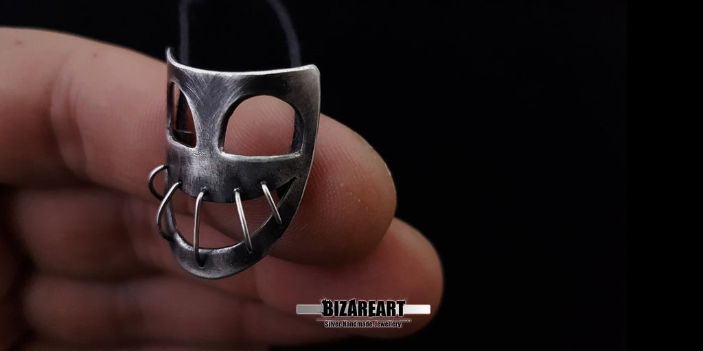 Macro close-up of the silver wire-stitched details and textured abrasions on the mask pendant held between fingers.