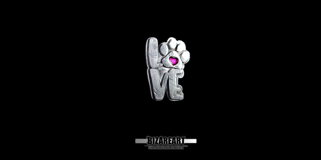 A high-resolution studio shot of a 3D sterling silver "LOVE" pendant against a solid black background. The letter "O" is replaced by a detailed dog paw print with a vibrant magenta heart at its center.