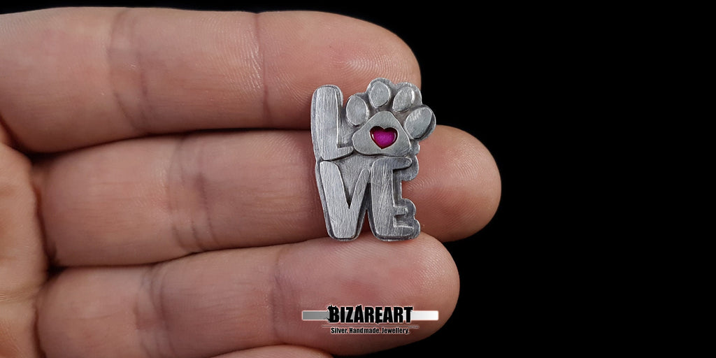 A top-down view of the pendant resting on a hand, showing the intricate swirling textures in the background and the precision of the magenta enamel inlay.