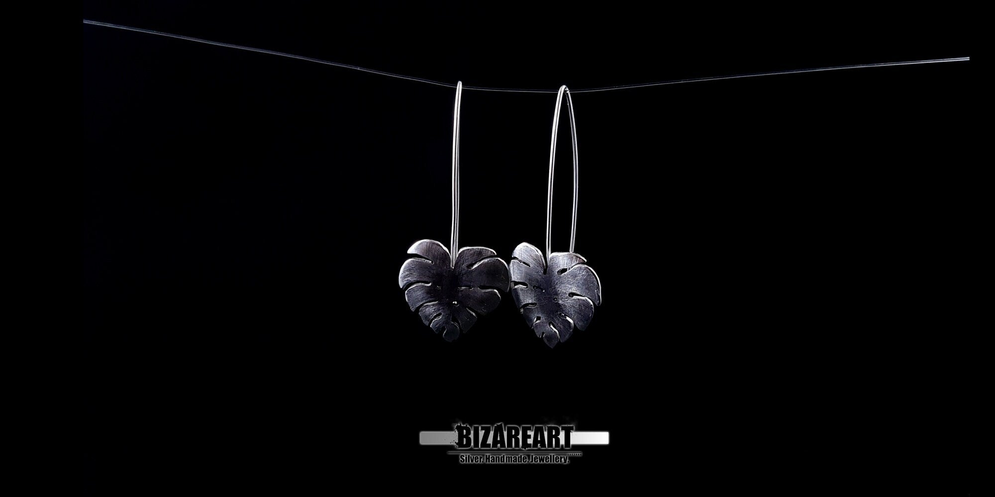 A pair of silver Monstera leaf earrings suspended from long wire hooks, showcasing their elegant silhouette and statement length.