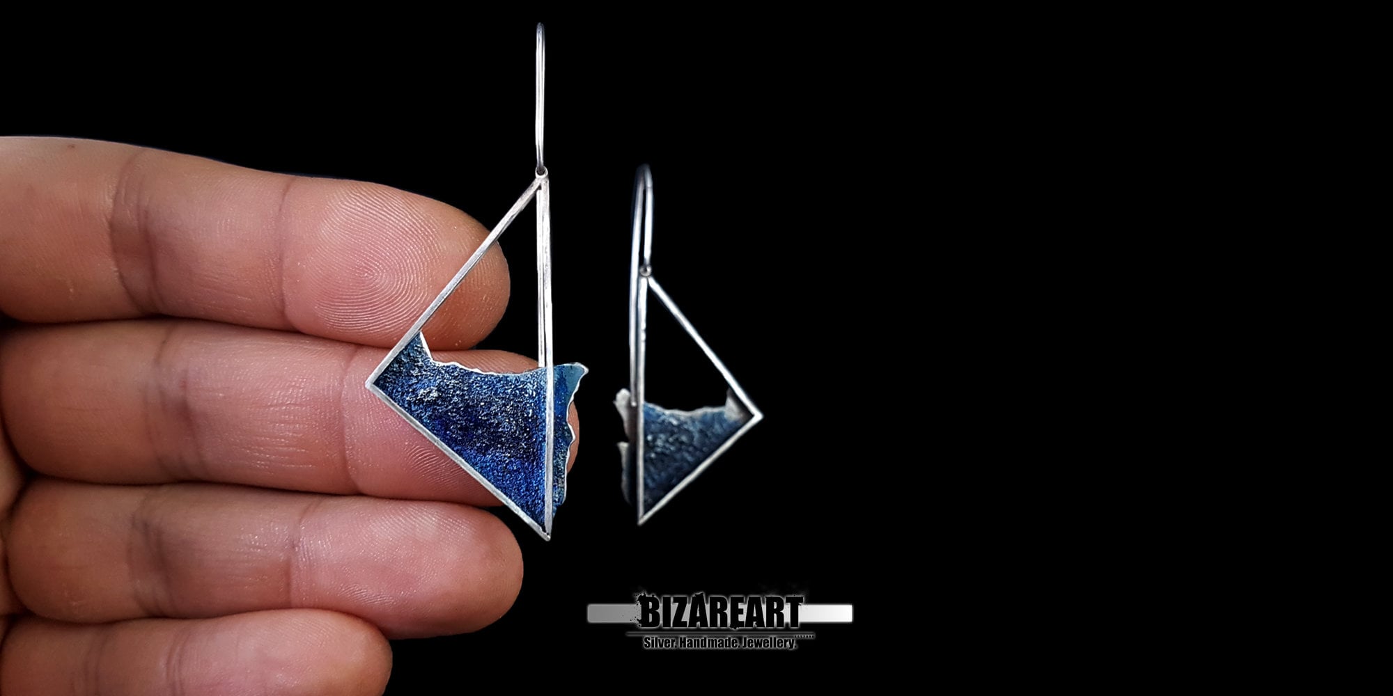 A hand holding one of the blue enamel silver triangle earrings to show the actual size, scale, and long wire hook length.