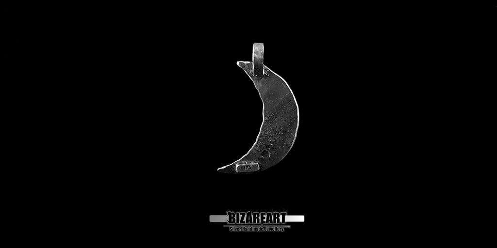 A studio product shot of the silver crescent moon pendant centered horizontally against a sharp black backdrop, showcasing its curved profile. A small white logo is visible beneath the piece.