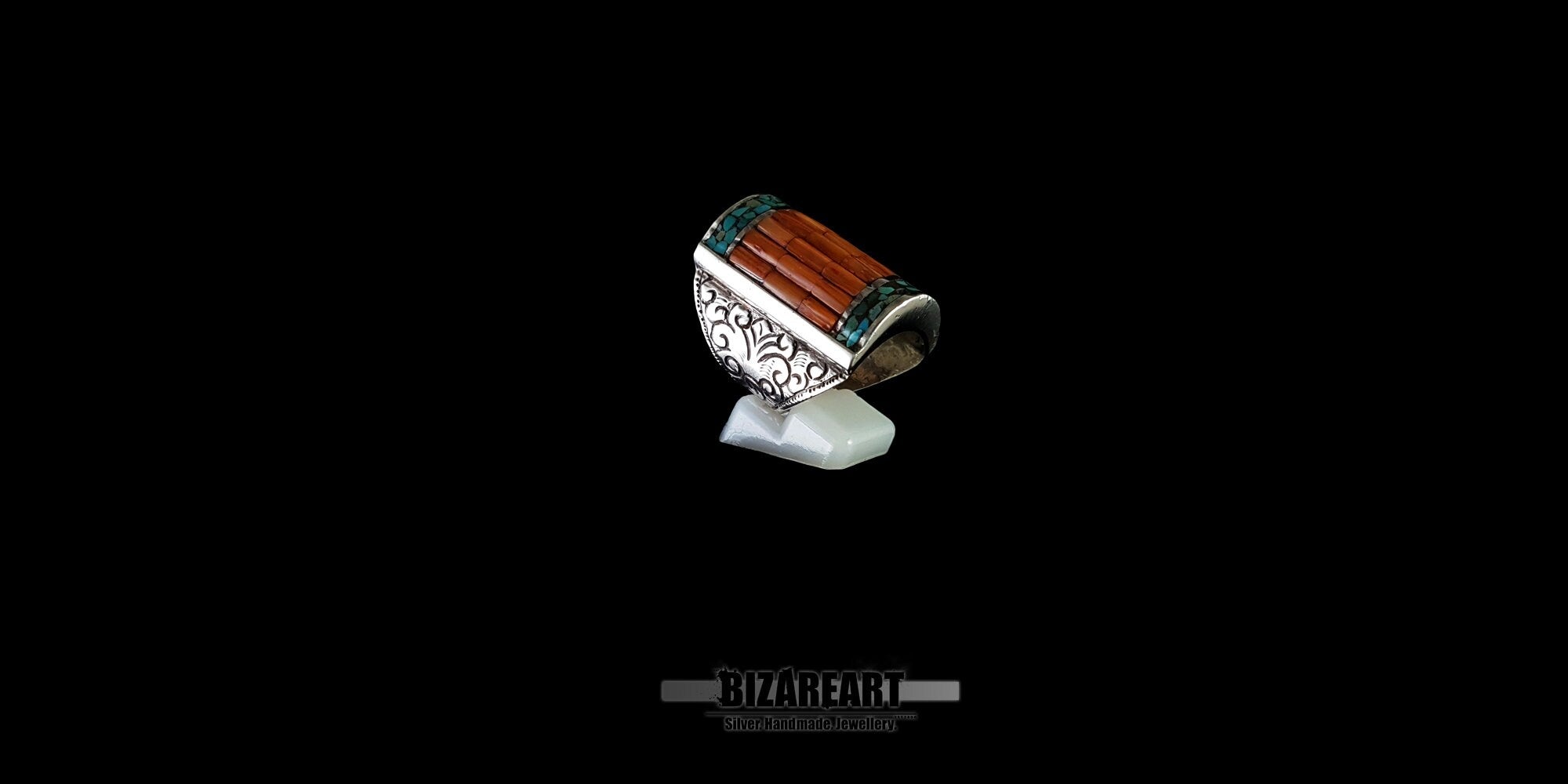 Side view of a vintage Tibeto-Nepalese silver statement ring, highlighting the detailed floral swirl engravings on the band and a vibrant red coral and turquoise mosaic inlay against a solid black background.