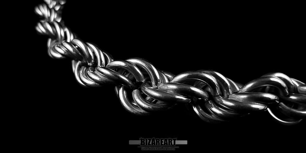 Side-angle macro shot of a silver rope chain on a black background, focusing on the depth and 3D architectural weave of the links.