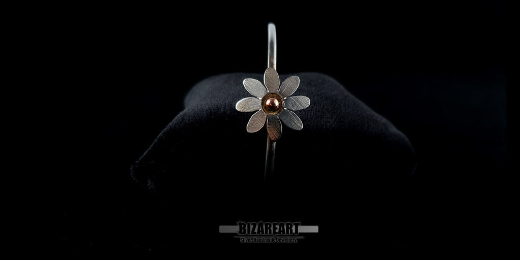 Front view of a handmade sterling silver daisy bangle bracelet with a copper pink gold center on a black jewelry cushion.