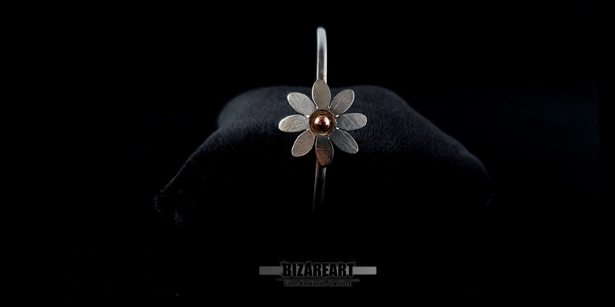 Front view of a handmade sterling silver daisy bangle bracelet with a copper pink gold center on a black jewelry cushion.