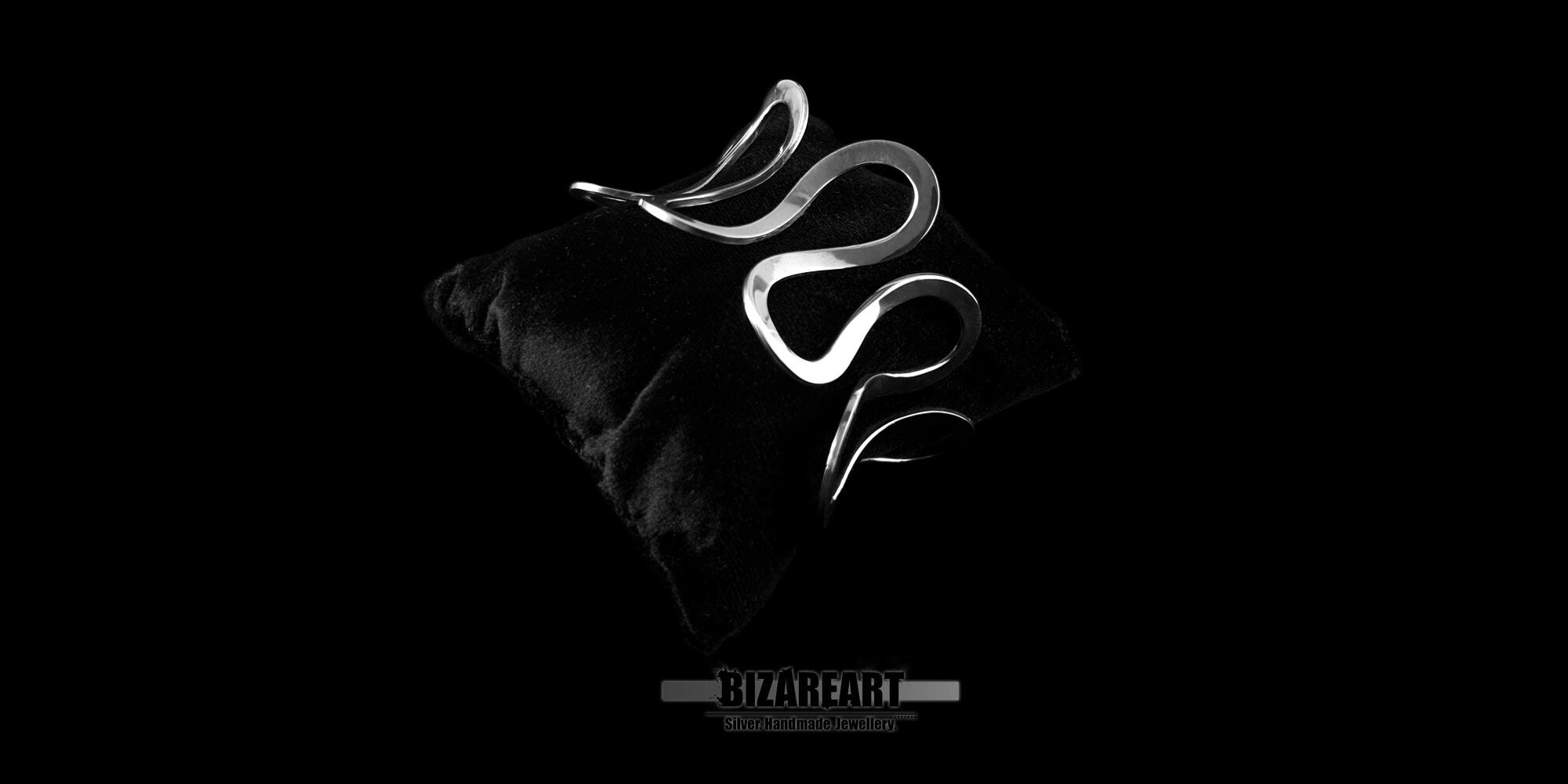 A product image featuring the same sculptural silver cuff bracelet resting dynamically atop a textured black fabric surface or cushion. The shot is presented in a wide aspect ratio with a stark black backdrop, highlighting the bracelet’s reflective metallic loops and elegant curves.