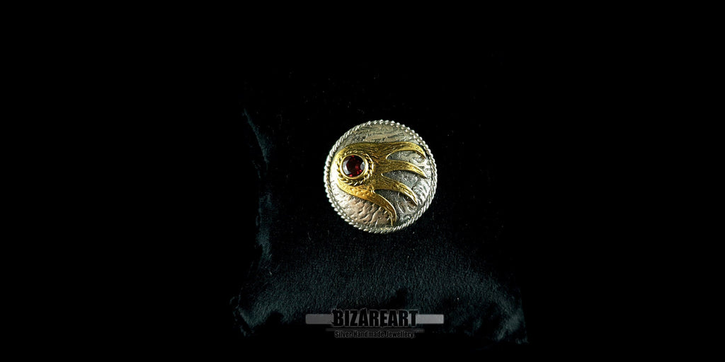 A high-contrast close-up of a round, handmade sterling silver pendant featuring a gold-gilded hand design and a central red garnet gemstone, set against a black background.