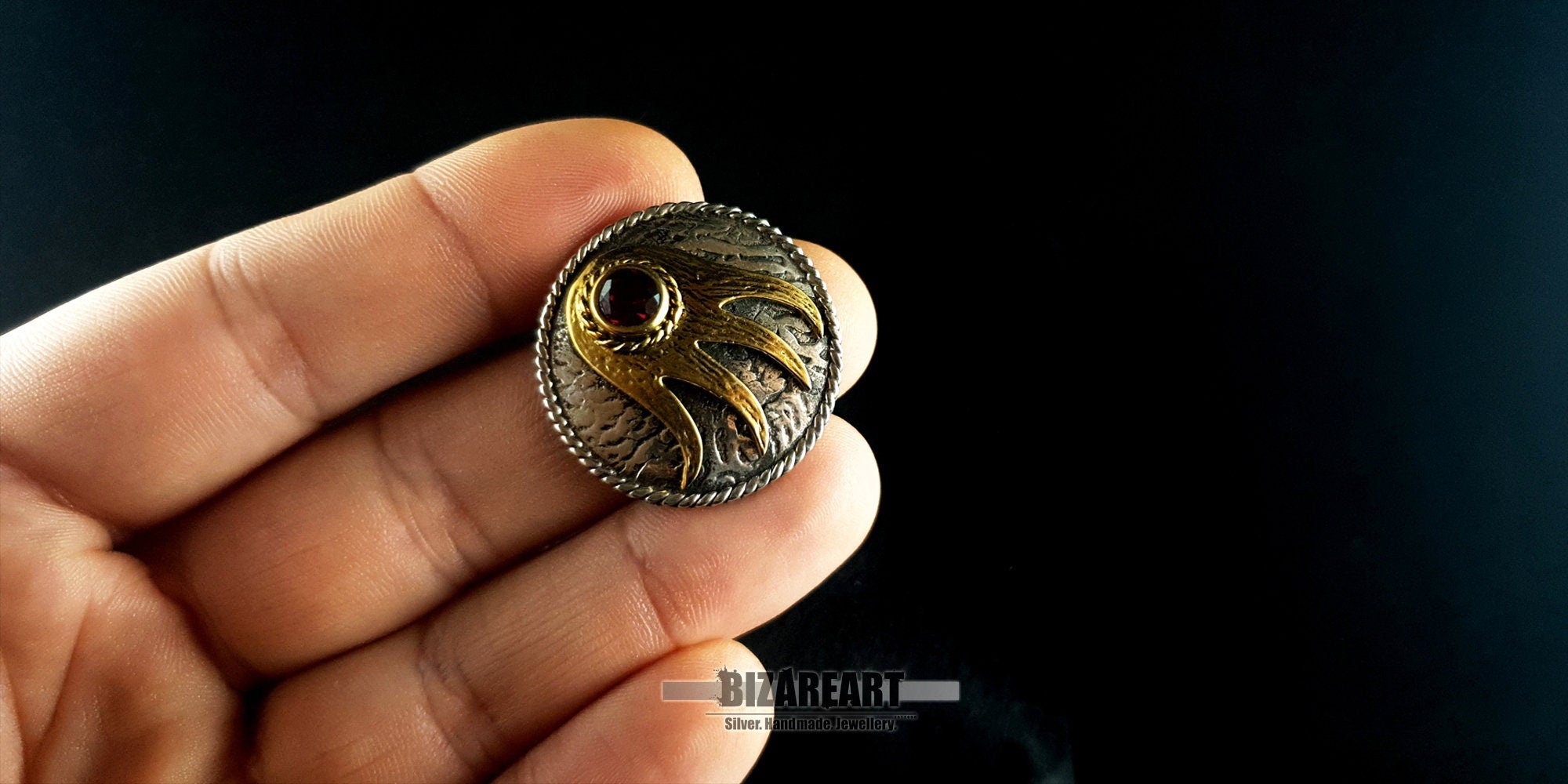 A person holding the small round silver and garnet pendant between their fingers to demonstrate its 3.0 cm scale and substantial 11.7g weight.
