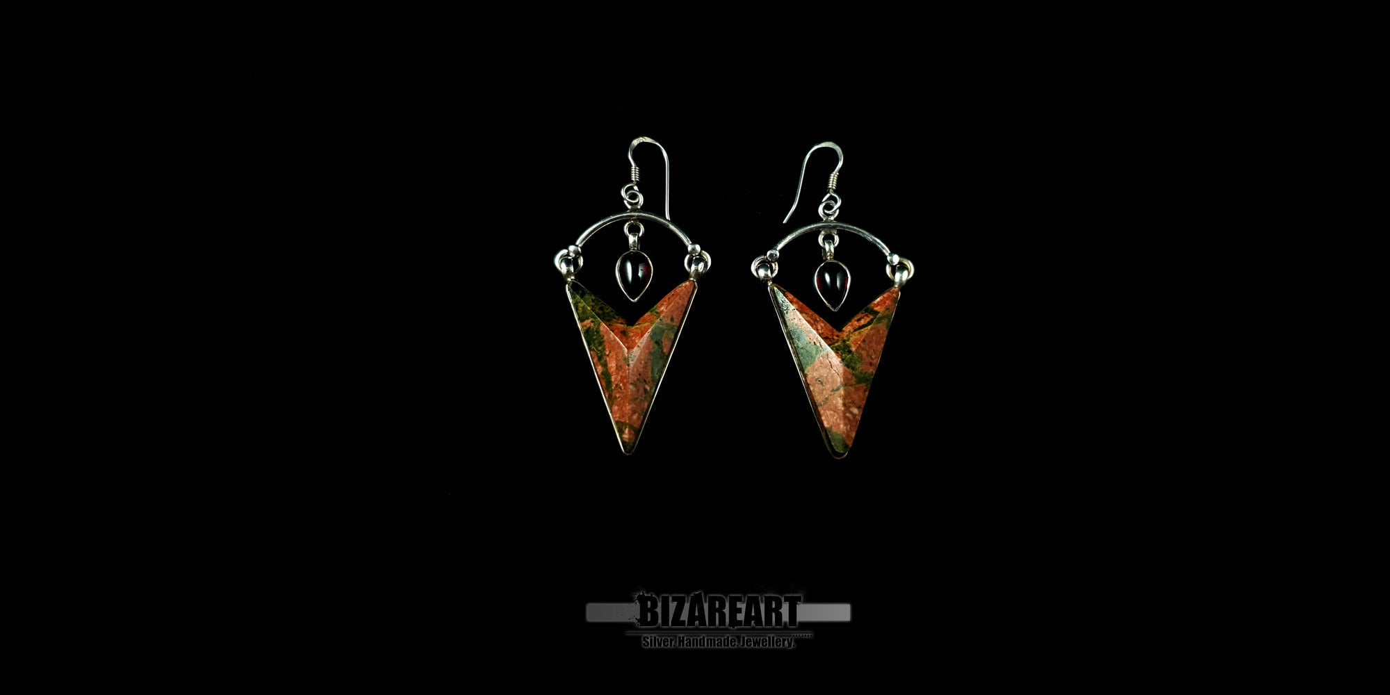 A pair of handmade Unakite earrings displayed centrally on a black background, showing the symmetrical design and the delicate silver arch holding the garnet stone.