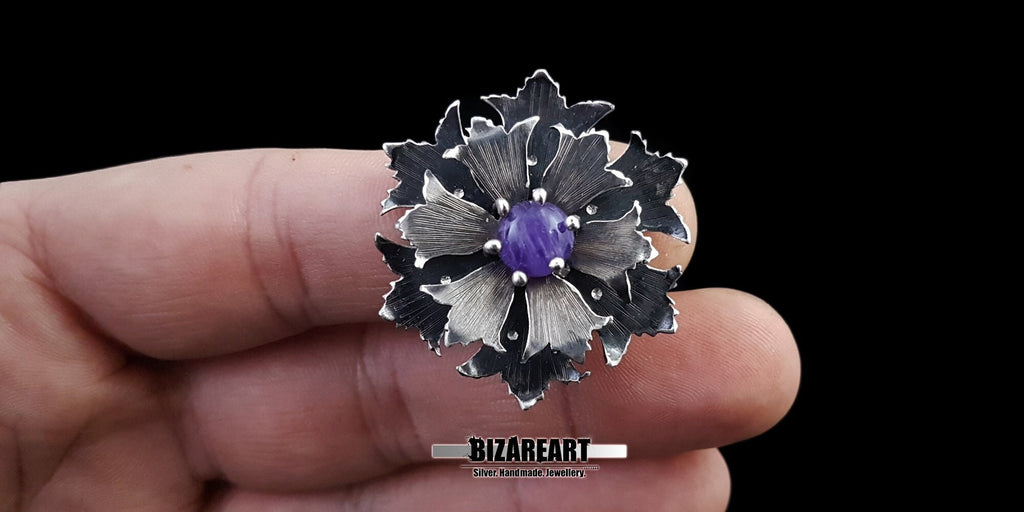 Close-up of a hand holding a large, intricate oxidized silver flower pendant with a vibrant purple amethyst center to show scale and detail.