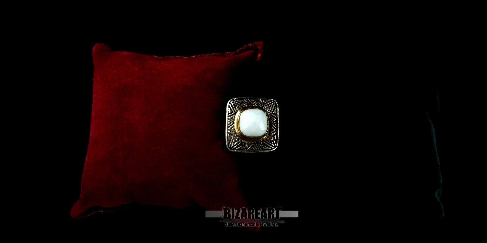 Top-down view of a handcrafted sterling silver statement ring featuring a square white agate stone with a gold-plated bezel, displayed next to a red velvet cushion on a black background.