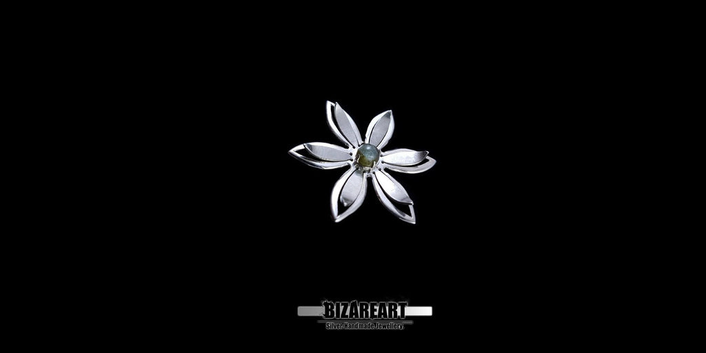 A high-contrast studio shot of a handcrafted silver flower pendant featuring six pointed petals and a central green gemstone, centered against a solid black background.