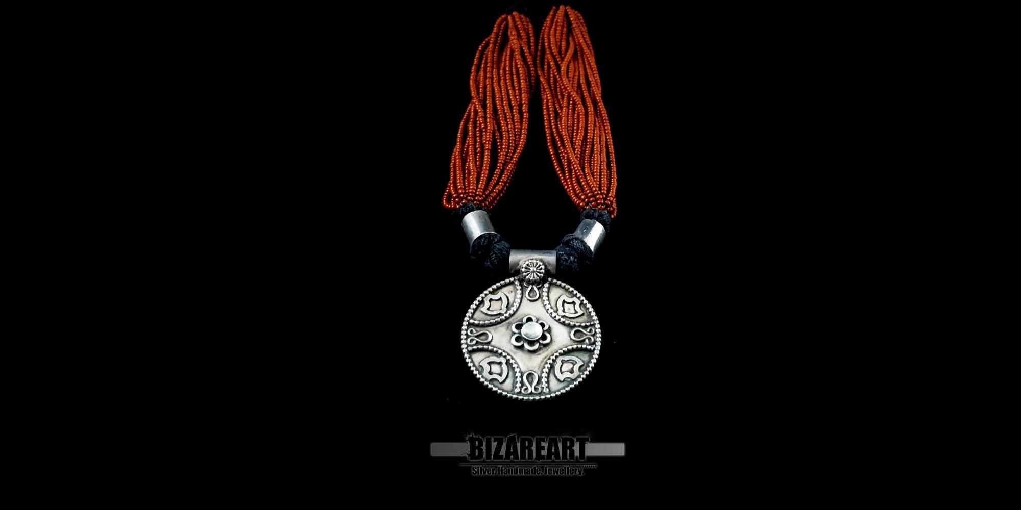 Front view of a handcrafted tribal necklace featuring multi-strand deep red garnet beads and a large, circular oxidized silver pendant with intricate floral engravings against a black background.