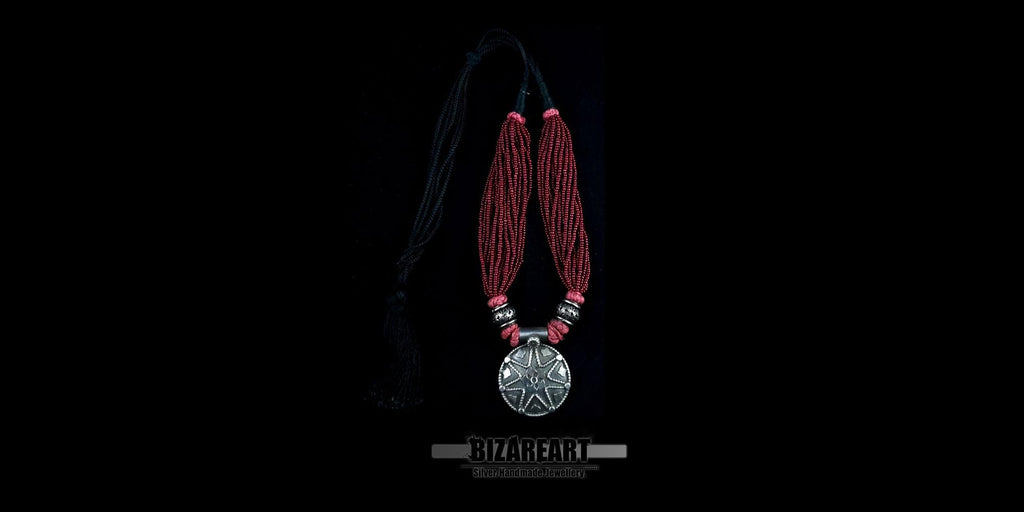Handcrafted multi-strand red bead necklace with a large round oxidized silver statement pendant, displayed flat on a black background.