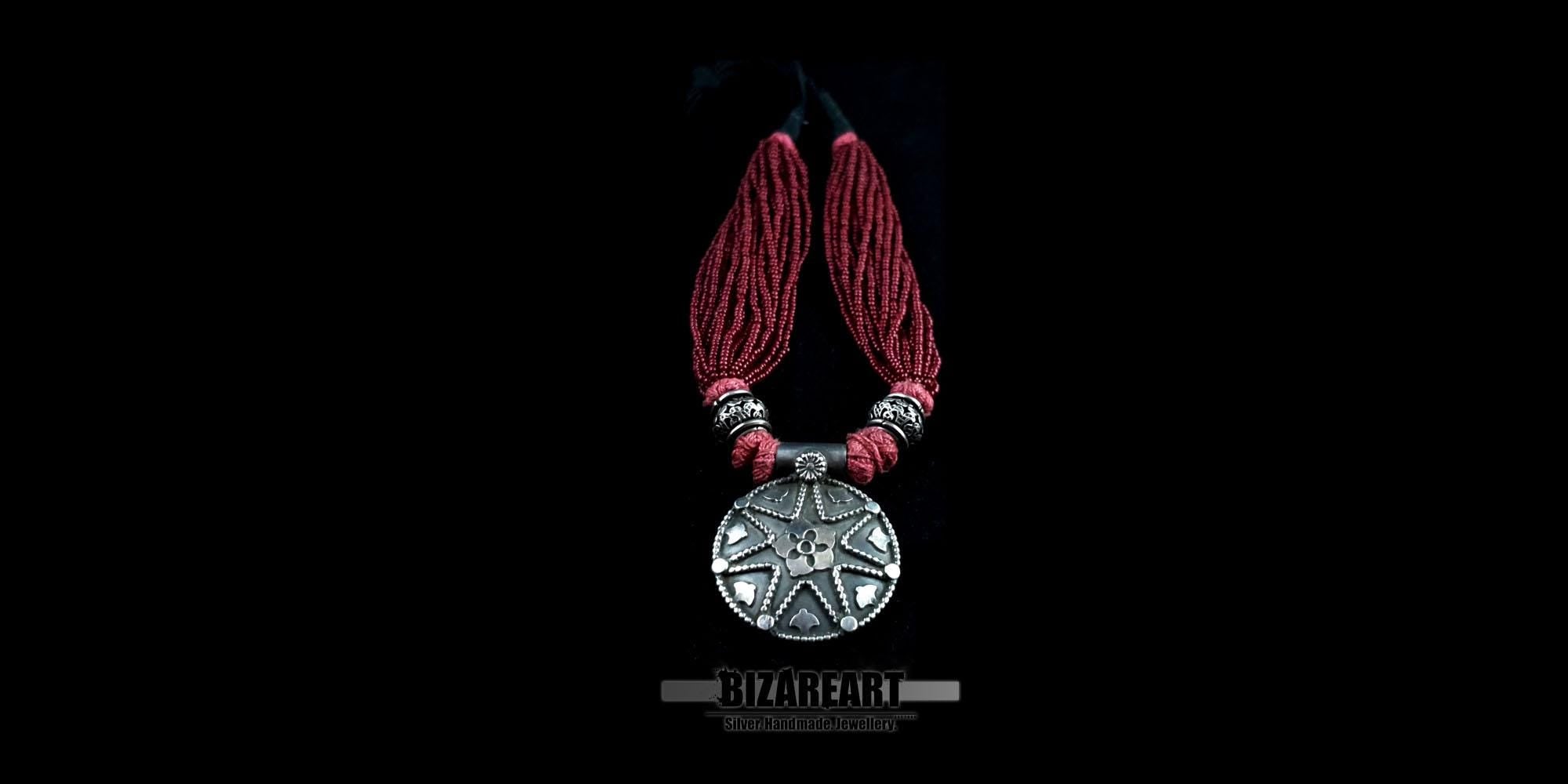 Close-up of a circular oxidized silver pendant featuring intricate floral and geometric details, attached to multiple strands of red seed beads.