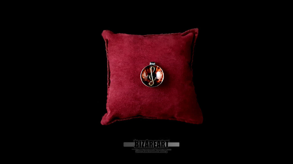 A top-down view of the handcrafted sterling silver and amber-toned enamel pendant resting on a plush, dark burgundy velvet display pillow against a black background.