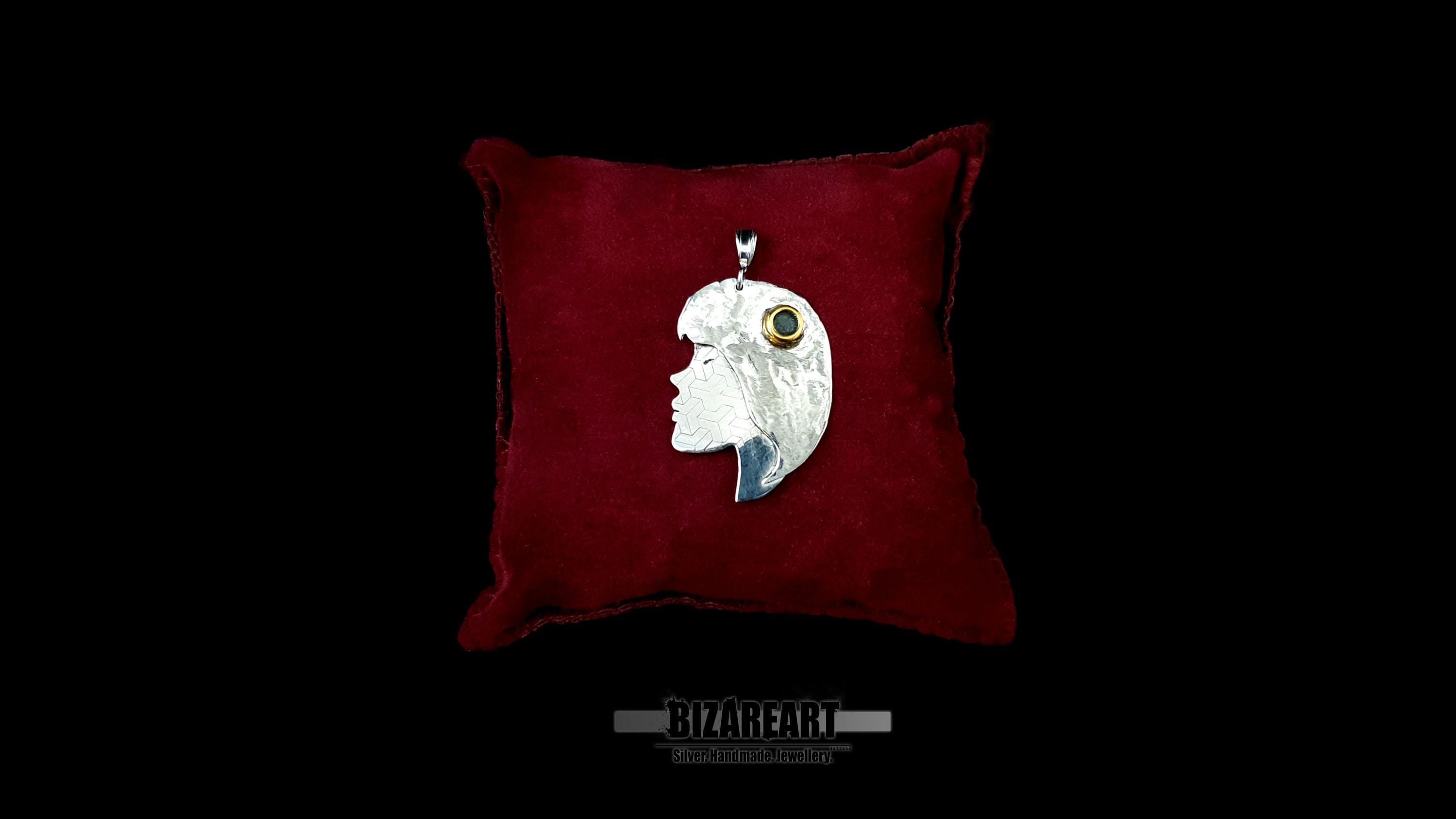 A profile-style sterling silver portrait pendant of a woman, featuring a gold-plated eye detail, resting on a plush dark red velvet cushion against a black background.