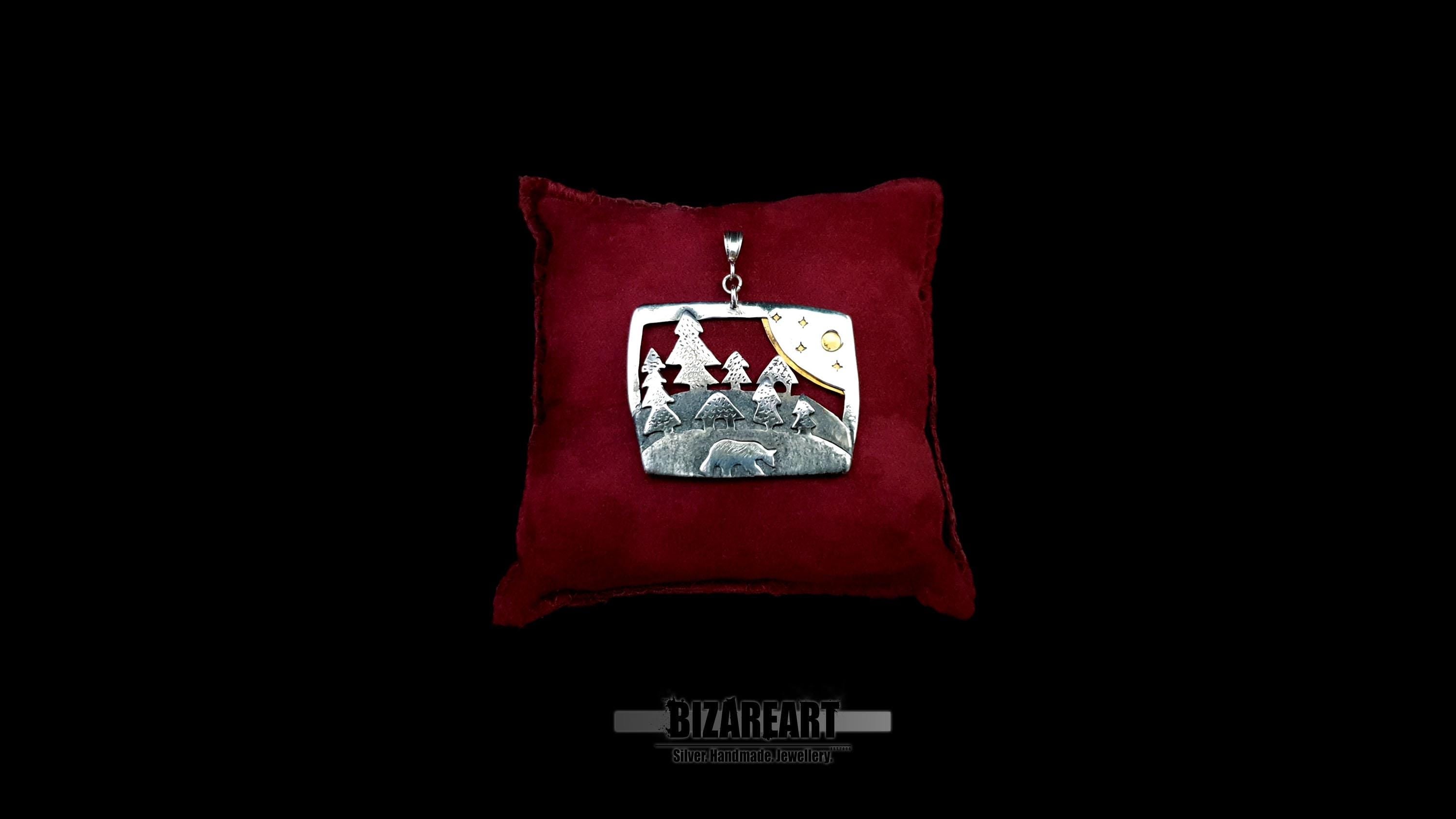 Handcrafted sterling silver bear and forest diorama pendant resting on a luxury crimson red velvet display pillow against a black background.