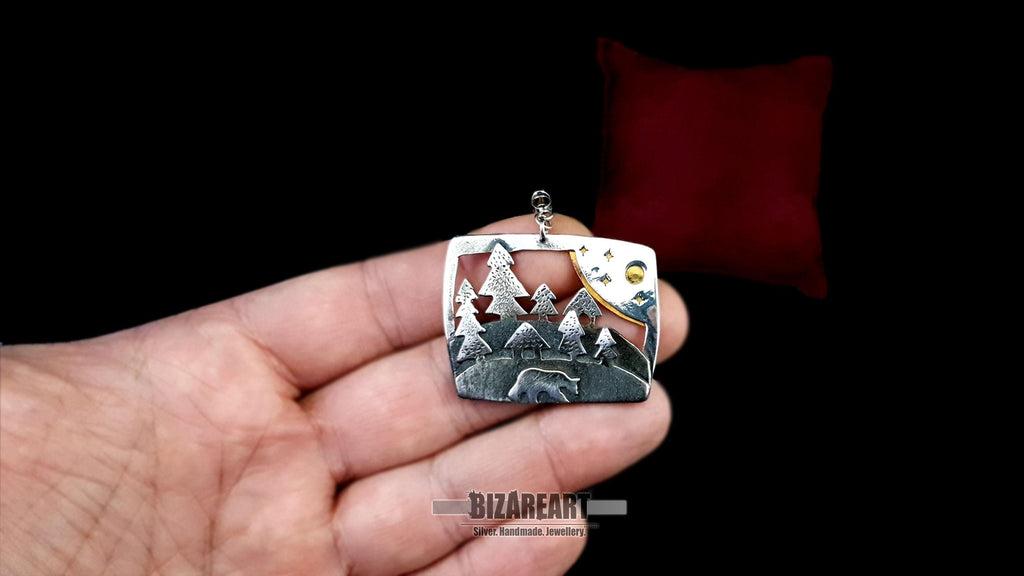 A person holding the artisan silver forest narrative pendant in their hand to show real-life size and scale, with a red display cushion in the background.
