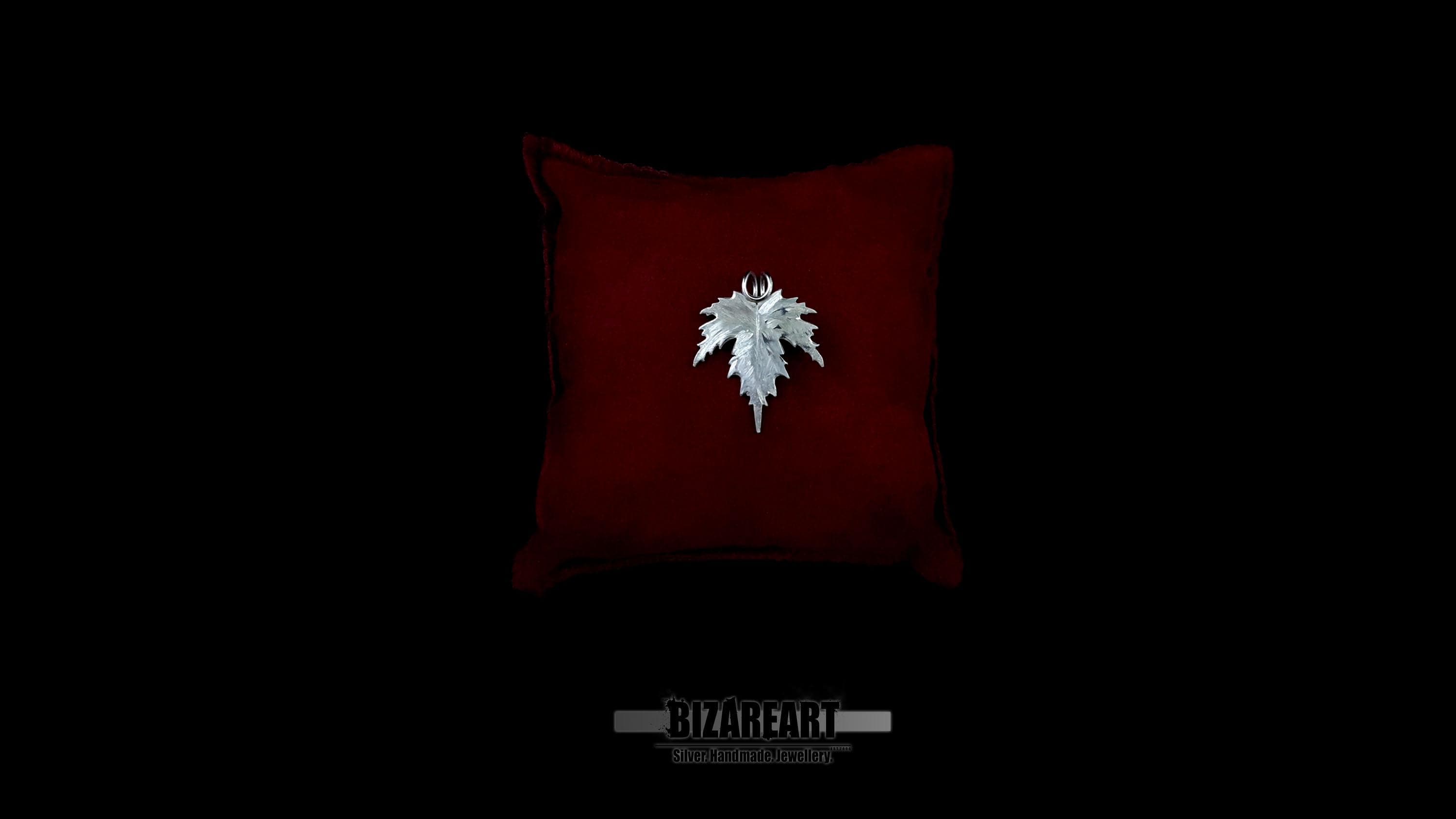 Top-down view of a hand-carved sterling silver leaf pendant resting on a plush red velvet cushion against a black background.