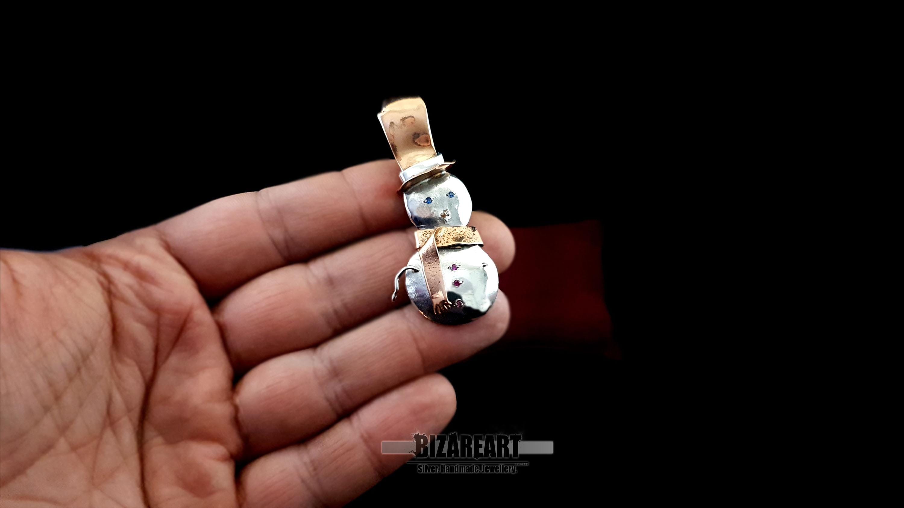A hand holding the snowman pendant to demonstrate its scale, showing the 6cm height and substantial 10g artisan build.