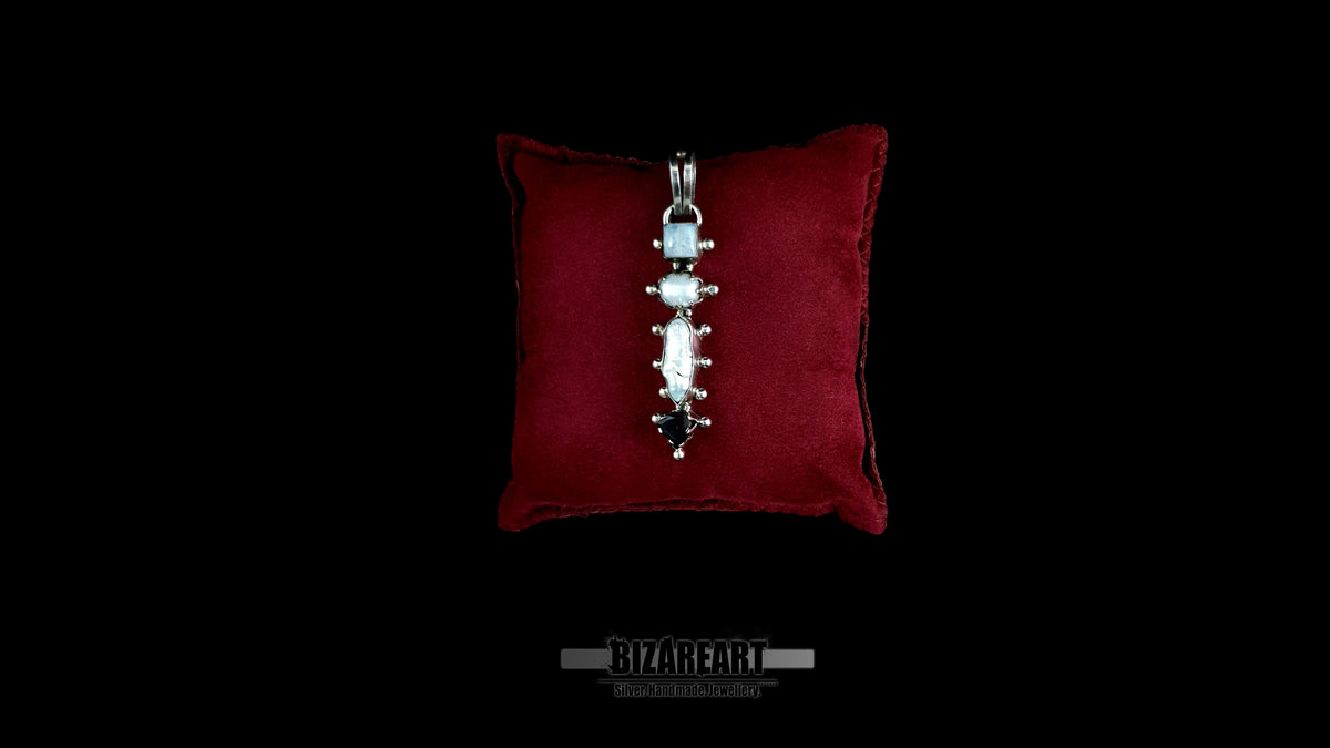 A detailed top-down view of the handcrafted sterling silver Celestial Quad Pendant, featuring four articulated settings with natural moonstone, pearl, and garnet, resting centrally on a plush maroon velvet presentation pillow against a black background.