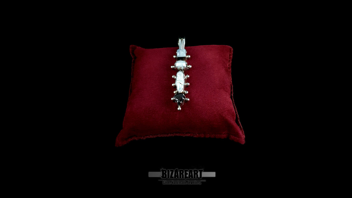 A high-angle studio shot of the sterling silver Celestial Quad Pendant, with its unique stack of four gemstones (moonstone, pearl, garnet), resting on a deep red velvet display cushion against a black backdrop.