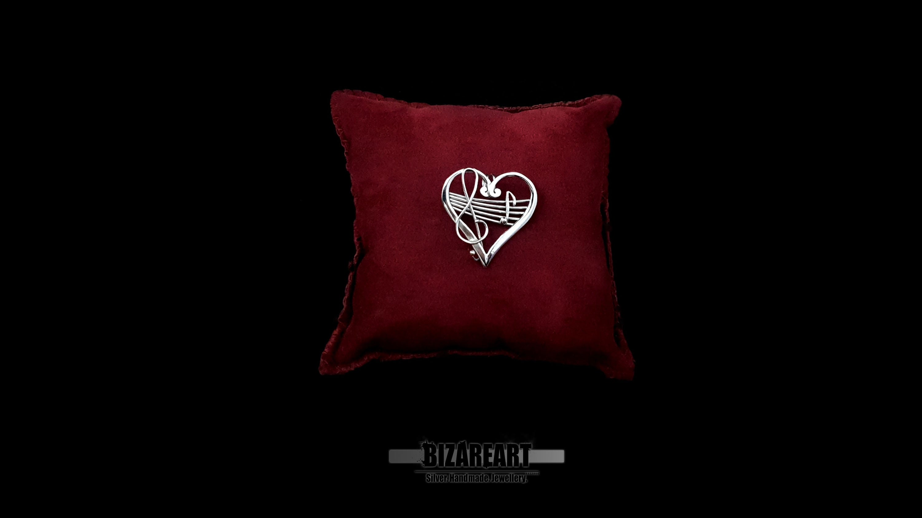 Hand-forged sterling silver heart melody pendant resting on a luxurious deep red velvet pillow, centered against a dramatic black background with a clean brand watermark below.