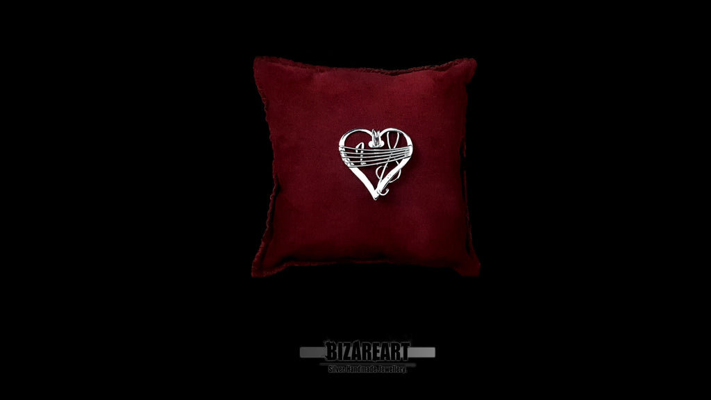 Front-facing product shot of the silver music-inspired heart pendant on a red velvet pillow, showcasing the intricate Sol clef and Quaver wire details.