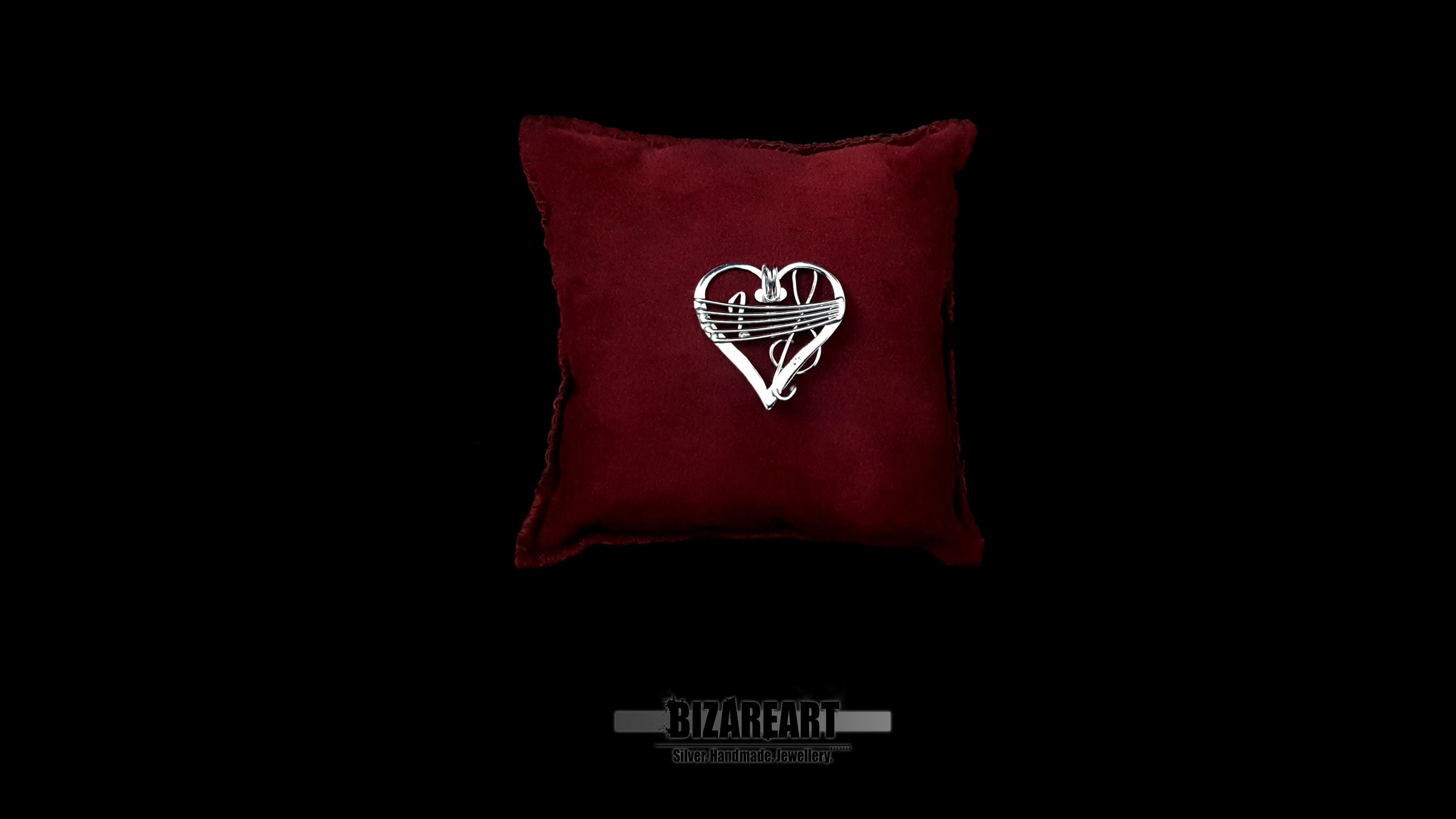 Front-facing product shot of the silver music-inspired heart pendant on a red velvet pillow, showcasing the intricate Sol clef and Quaver wire details.