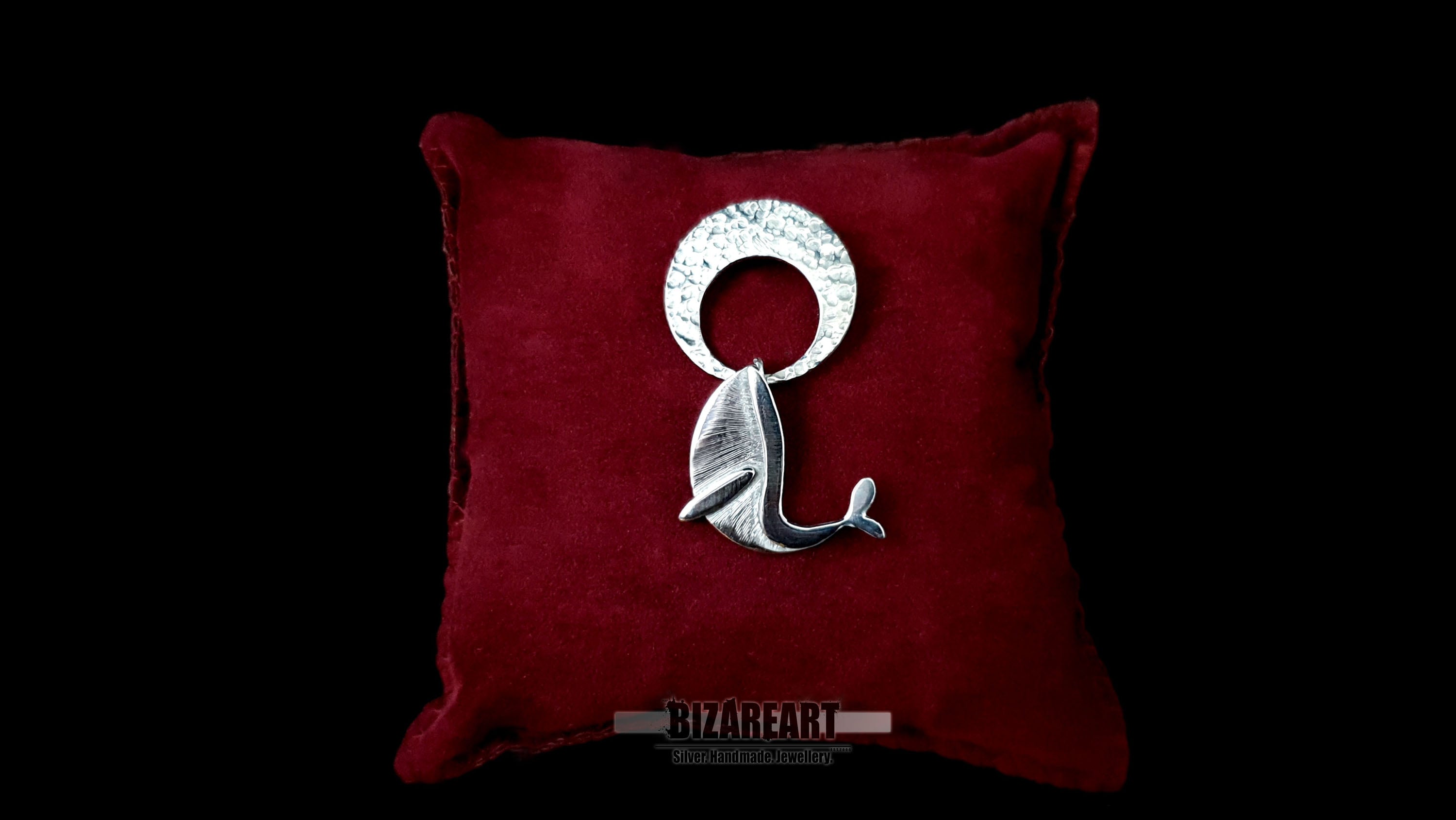 A handmade sterling silver whale and moon pendant displayed on a luxury dark red velvet pillow. The artisan jewelry piece showcases a hammered silver crescent moon and a detailed, hand-engraved whale breaching beneath it against a black background.