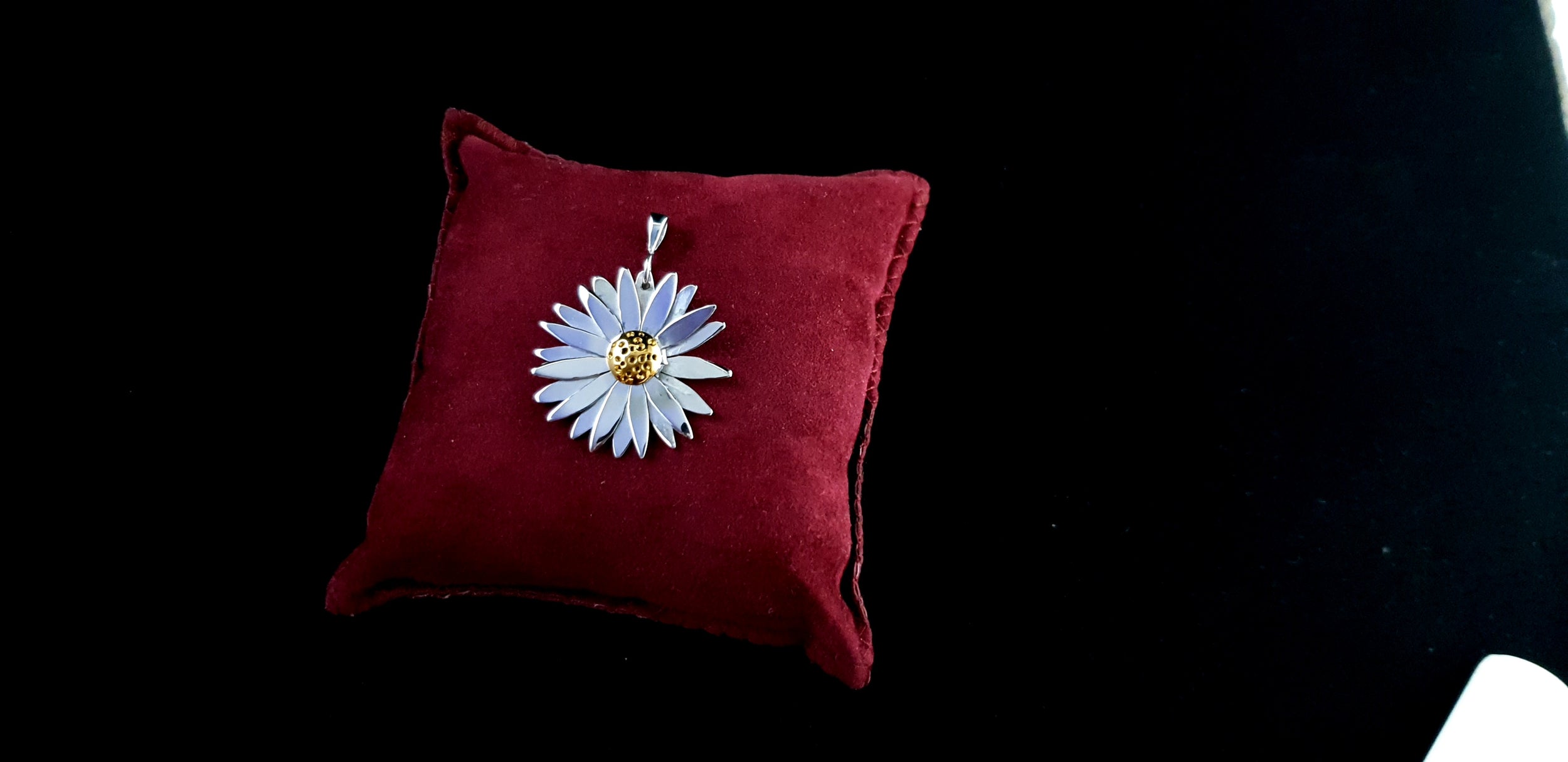 A video preview showing the sterling silver daisy pendant resting on a red velvet pillow, demonstrating how the light reflects off the polished petals and gold-plated center.