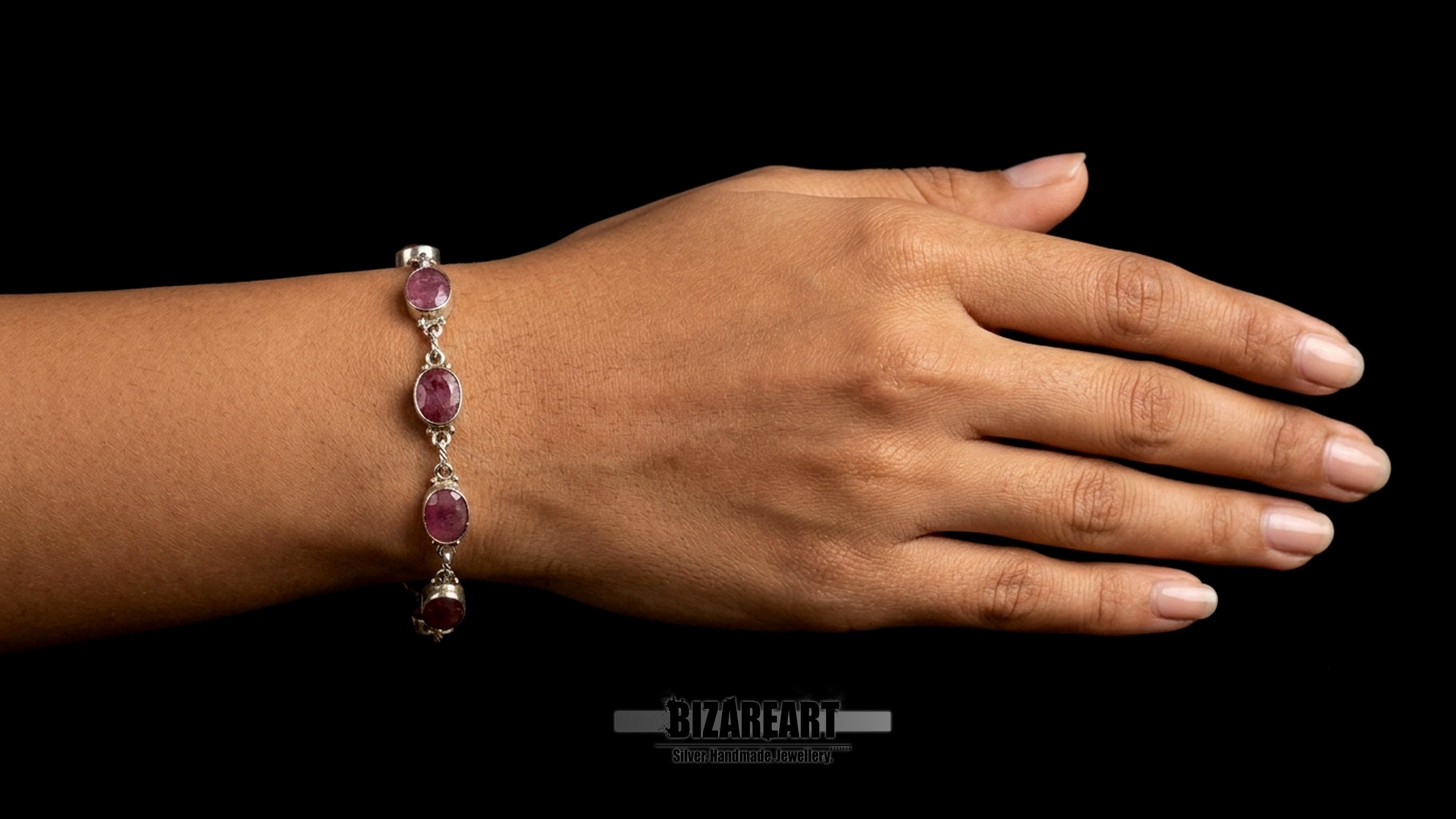 Close-up on-model shot of a woman's wrist wearing the handmade sterling silver link bracelet with six raw oval rubies, presented against a solid black studio background.