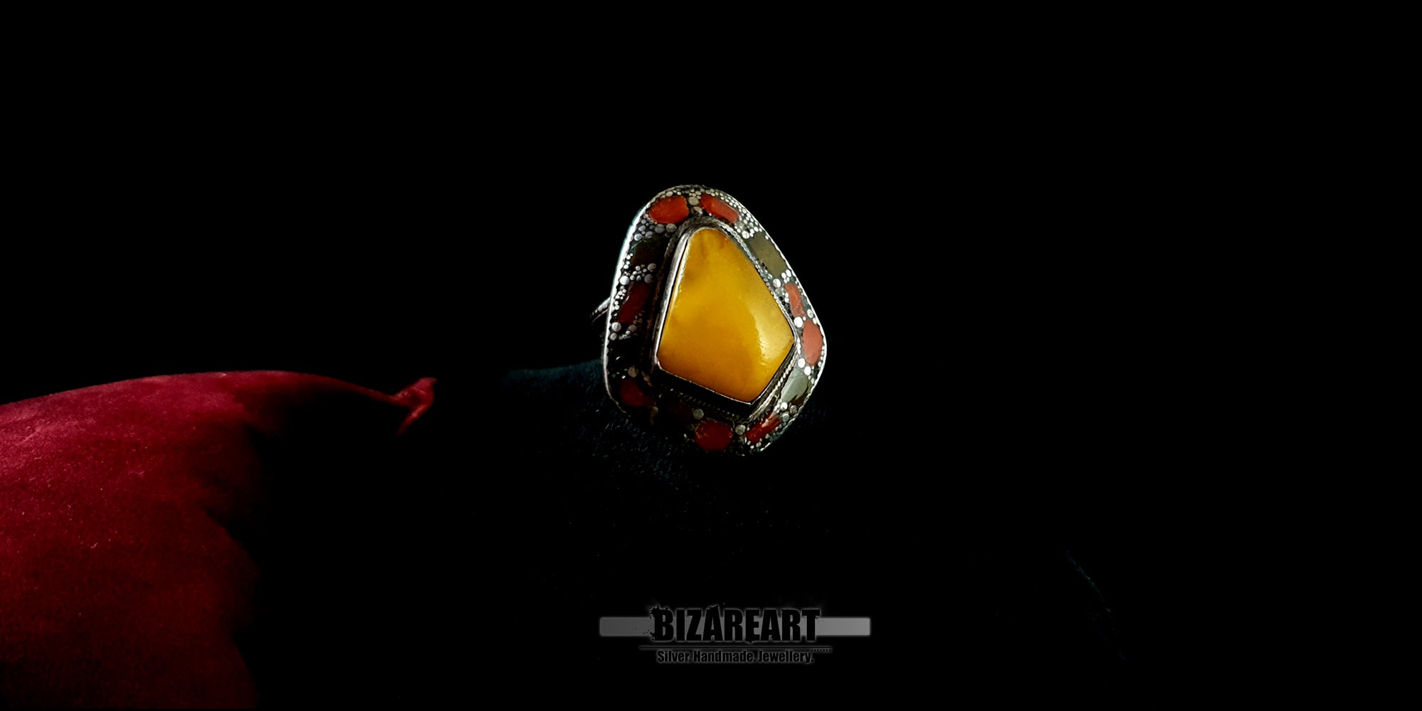 Large teardrop-shaped yellow amber ring with red coral accents in an engraved silver setting, resting on a red velvet pillow by BIZAREART.