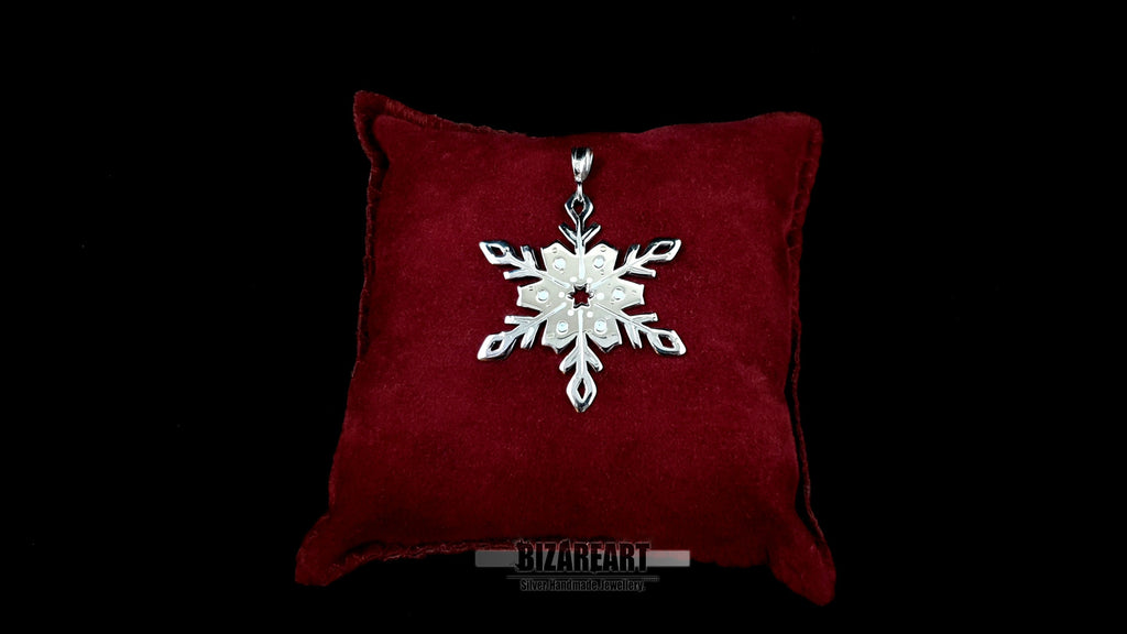 A hand-pierced silver snowflake pendant with iridescent enamel "melting" droplets, displayed on a plush crimson velvet jewelry cushion against a dark background.
